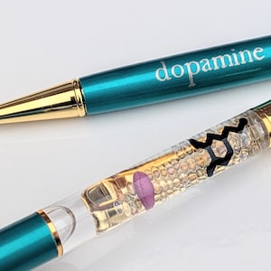 May include: Two teal pens with gold accents. One pen has the word "dopamine" printed on it. The other pen has a clear barrel with a purple and black design inside. The pens are on a white surface.