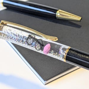 May include: Two pens on a black notebook. One pen is black with a gold tip. The other pen is clear with a gold clip and contains small objects, including a pink eraser and tiny black glasses.