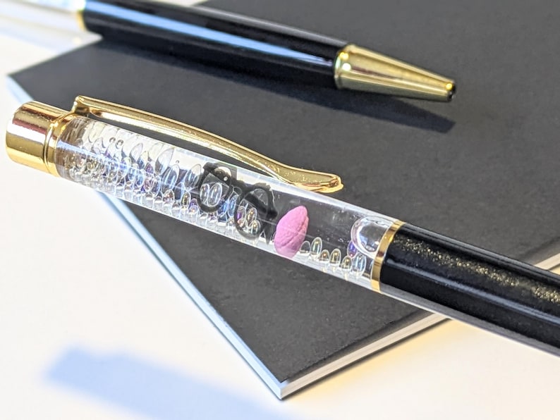 May include: Two pens on a black notebook. One pen is black with a gold tip. The other pen is clear with a gold clip and contains small objects, including a pink eraser and tiny black glasses.