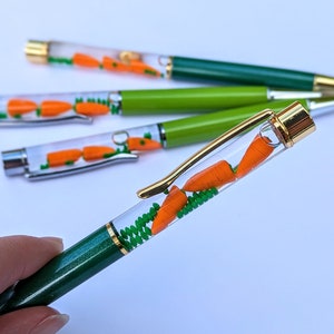 May include: A collection of novelty pens with a carrot theme. Each pen features a clear barrel filled with miniature orange carrots and green foliage. The pens have green or lime green barrels with gold or silver accents.