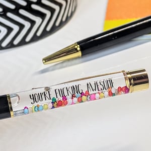 May include: Two black pens with gold detailing. One pen features the text "YOU'RE FUCKING AWESOME" in black, with colourful beads inside the clear barrel. The other pen is plain.
