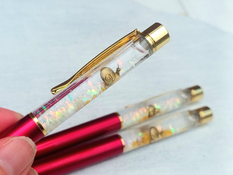 May include: Three red and gold pens with clear barrels filled with iridescent glitter and a gold snail charm.