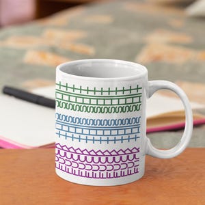 May include: White ceramic mug featuring a repeating pattern of colorful geometric designs in green, blue, and purple. The mug has a curved handle and is sitting on a wooden surface.