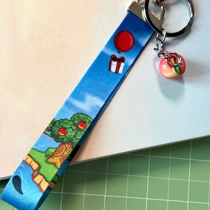 Könnte beinhalten: Ein blaues Schlüsselband mit einem Cartoon-Landschaftsdesign mit einem roten Ballon, einem Geschenk und einem Baum mit roten Äpfeln. Ein Pfirsich-Anhänger hängt am silbernen Schlüsselring. Das Band besteht aus strapazierfähigem Stoff.