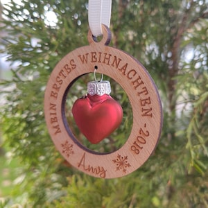 Puede incluir: Adorno navideño de madera con una bola roja en forma de corazón dentro de un marco circular. El marco está grabado con las palabras "MEIN ERSTES WEIHNACHTEN - 2018" y el nombre "Amy". El adorno cuelga de una cinta blanca.
