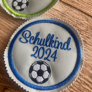 Könnte beinhalten: Ein weißer Stoff-Patch mit blauem Rand und einem Fußball-Design. Der Patch trägt die Aufschrift "Schulkid 2024" in blauer Kursivschrift.