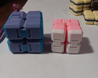 Könnte beinhalten: Zwei Fidget-Spielzeuge werden gezeigt. Eines ist ein blau-grauer Würfel mit türkisfarbenen Akzenten. Das andere ist ein rosa-weißer Würfel. Im Hintergrund befinden sich mehrere andere ähnliche Spielzeuge, darunter gelbe und schwarze.
