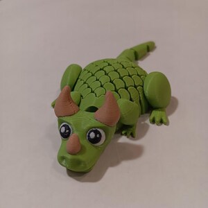 Peut inclure: Figurine de dragon vert imprimée en 3D avec un corps texturé et détaillé. Le dragon a de grands yeux ronds, un museau brun clair et des cornes assorties. La queue et les membres du dragon sont également visibles.