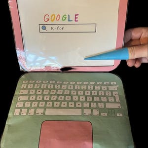 以下が含まれることがあります： キーボードと画面を備えた、ピンクと緑のラップトップ型のクラフト。画面にはGoogleのロゴと「K-POP」と入力された検索バーが表示されています。青いポインターが画面に触れています。