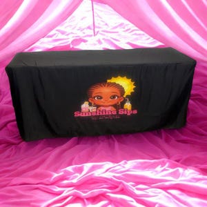 May include: A black table cover featuring the "Sunshine Sips" logo with a cartoon girl and a sun graphic. The cover is set against a backdrop of pink fabric, creating a vibrant contrast.