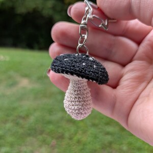 May include: A handmade crochet mushroom keychain. The mushroom cap is black with white spots, and the stem is beige. The keychain has a silver-colored clasp.