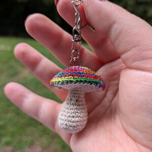 May include: A handmade crochet mushroom keychain. The mushroom has a rainbow-colored cap and a beige stem. The keychain is attached to a silver-colored clasp.
