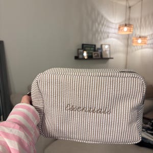 May include: A rectangular, seersucker-striped cosmetic bag in brown and white. The word "Essentials" is embroidered in a cursive font on the front of the bag. The bag is held by a person wearing a pink and white striped sleeve.