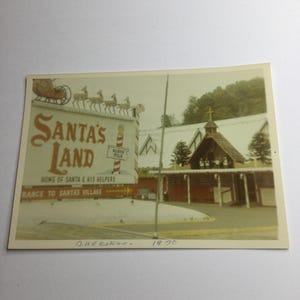 May include: Vintage photograph of Santa's Land, a Christmas-themed attraction. The sign reads "Santa's Land" with a depiction of Santa's sleigh and reindeer. The sign also indicates the North Pole and the entrance to Santa's Village. The photo was taken in Cherokee in 1970.