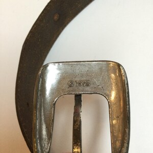 Może przedstawiać: Zbliżenie srebrnej klamry paska o prostokątnym kształcie i centralnym drążku. Klamra ma zużyty wygląd, widoczny jest napis "&copy;1998". Na zdjęciu widoczny jest r&oacute;wnież fragment brązowego sk&oacute;rzanego paska.