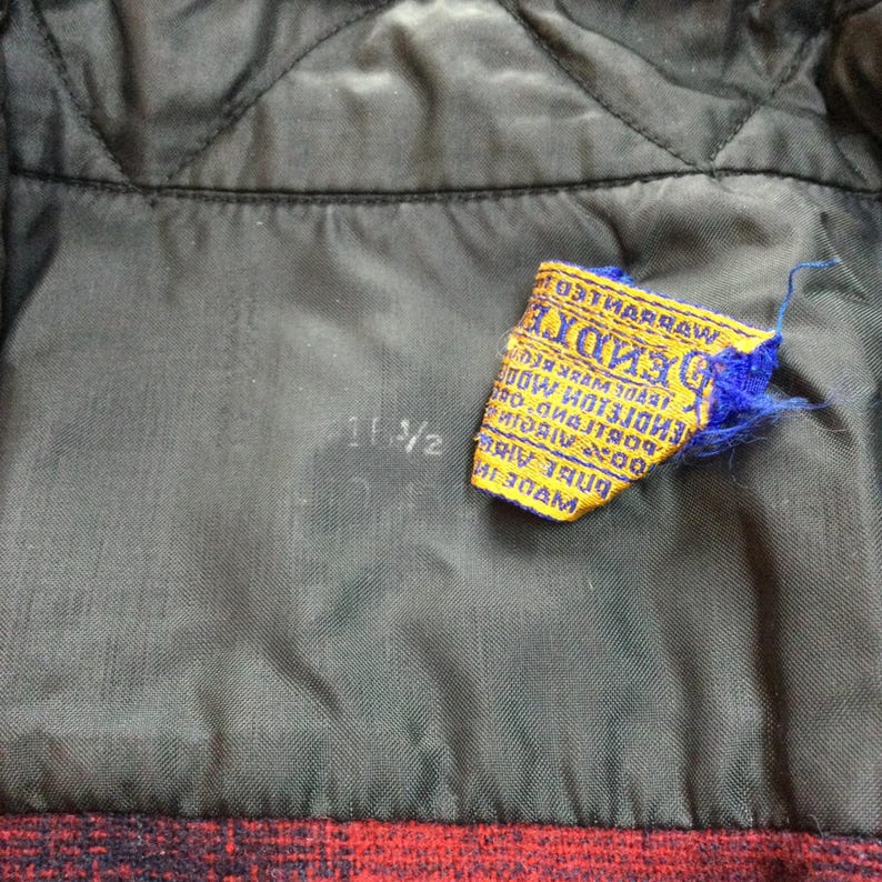 May include: Close-up of the inside lining of a garment. The lining is dark gray with a quilted pattern. A yellow and blue tag with text is attached. The measurement "11 1/2" is printed on the lining.