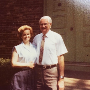 May include: A vintage photograph of a smiling couple standing together outdoors. The man wears a white short-sleeved shirt, brown pants, and a patterned tie. The woman wears a light blue top and a dark skirt. A brick wall and green door are in the background.