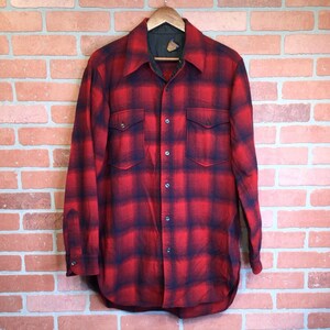 May include: A red and black plaid flannel shirt with two chest pockets and a button-down front. The shirt is hanging on a wooden hanger against a brick wall background. The shirt has a classic, vintage style.