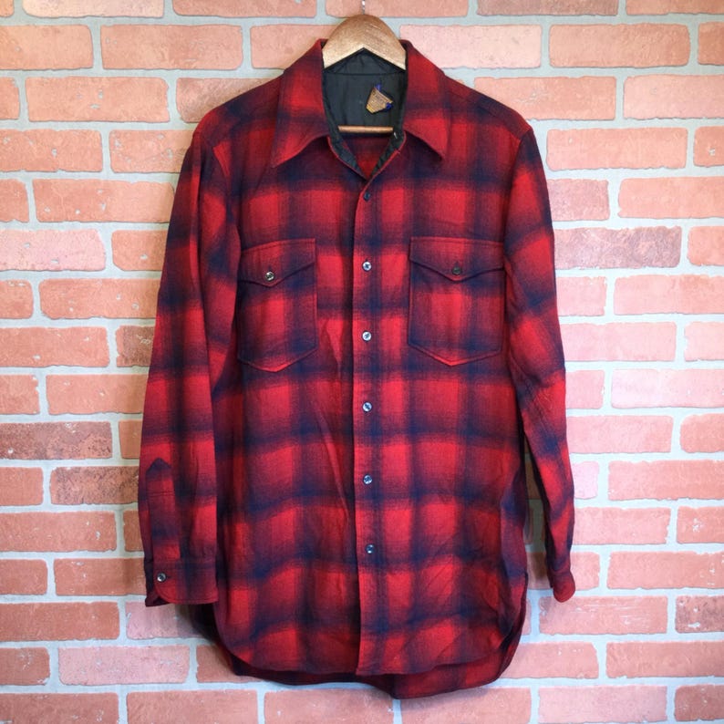 May include: A red and black plaid flannel shirt with two chest pockets and a button-down front. The shirt is hanging on a wooden hanger against a brick wall background. The shirt has a classic, vintage style.
