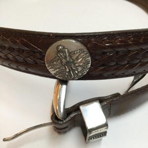 Może przedstawiać: Brązowy sk&oacute;rzany pasek z plecionym wzorem i srebrną klamrą. Pasek posiada okrągłą srebrną emalię z golfistą w akcji. Pasek ma szerokość około 3,8 cm.