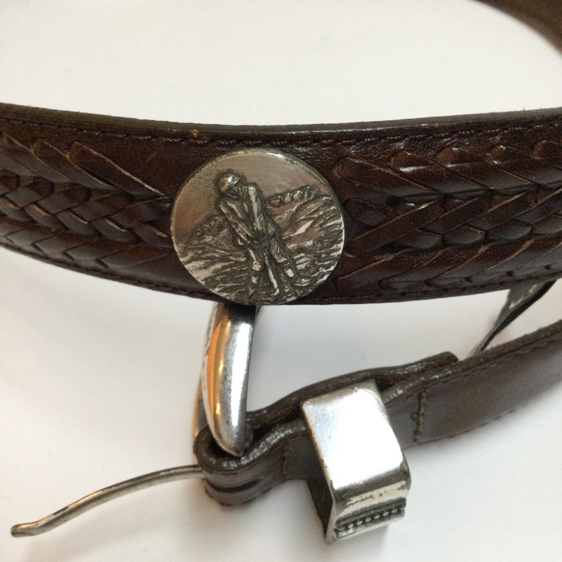 Może przedstawiać: Brązowy sk&oacute;rzany pasek z plecionym wzorem i srebrną klamrą. Pasek posiada okrągłą srebrną emalię z golfistą w akcji. Pasek ma szerokość około 3,8 cm.