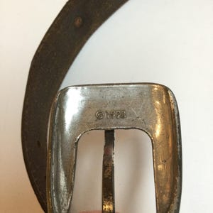 Może przedstawiać: Zbliżenie srebrnej klamry paska o prostokątnym kształcie i centralnym drążku. Klamra ma wygrawerowany w metalu napis "&copy;1998". Częściowo widoczny jest brązowy sk&oacute;rzany pasek.