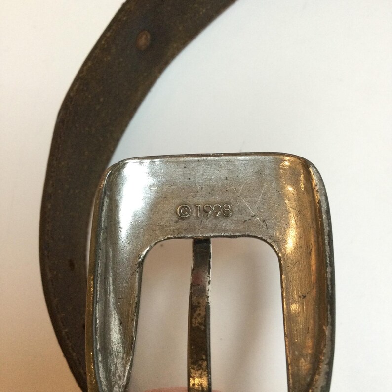 Może przedstawiać: Zbliżenie srebrnej klamry paska o prostokątnym kształcie i centralnym drążku. Klamra ma wygrawerowany w metalu napis "&copy;1998". Częściowo widoczny jest brązowy sk&oacute;rzany pasek.