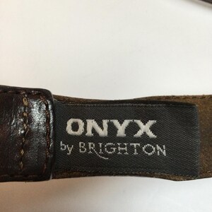 Może przedstawiać: Zbliżenie brązowego sk&oacute;rzanego paska z nazwą marki "ONYX by BRIGHTON" wydrukowaną na białym tle na ciemnoszarej etykiecie. Pasek ma bogaty, ciemnobrązowy kolor z widocznymi szwami.