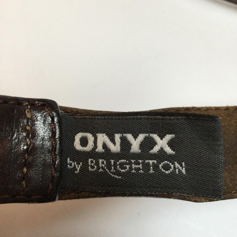 Może przedstawiać: Zbliżenie brązowego sk&oacute;rzanego paska z nazwą marki "ONYX by BRIGHTON" wydrukowaną na białym tle na ciemnoszarej etykiecie. Pasek ma bogaty, ciemnobrązowy kolor z widocznymi szwami.
