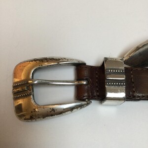 Może przedstawiać: Zbliżenie brązowego sk&oacute;rzanego paska ze srebrną klamrą. Klamra ma dekoracyjny wz&oacute;r i nosi ślady zużycia. Pasek to klasyczny dodatek.