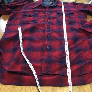 May include: A red and black plaid flannel shirt with two chest pockets and a button-down front. The shirt is being measured with a tape measure. The shirt's length is approximately 34 inches.