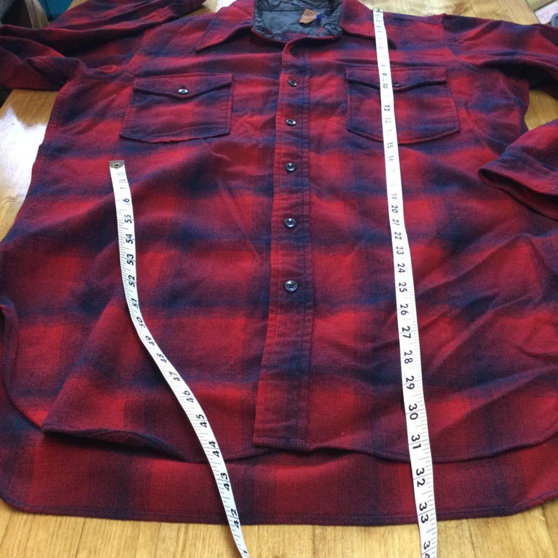 May include: A red and black plaid flannel shirt with two chest pockets and a button-down front. The shirt is being measured with a tape measure. The shirt's length is approximately 34 inches.