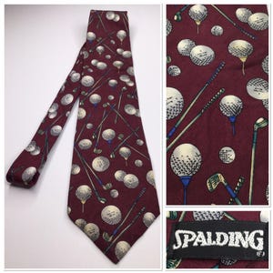 May include: A burgundy tie featuring a repeating pattern of golf balls and golf clubs. The golf balls are white with black detailing, and the clubs are green and blue. The tie has a Spalding label.