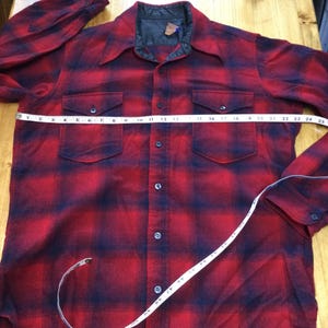 May include: Red and black plaid flannel shirt with two chest pockets and a button-down front. The shirt is being measured with a tape measure, showing a chest measurement of approximately 25 inches.
