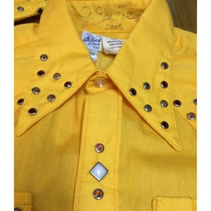 Può includere: Una camicia a maniche lunghe giallo brillante con colletto a punta decorato con borchie color argento. L'abbottonatura frontale presenta un bottone a pressione centrale bianco a forma di diamante affiancato da due bottoni metallici.