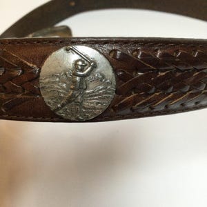 Może przedstawiać: Brązowy sk&oacute;rzany pasek z plecionym wzorem i srebrną okrągłą klamrą. Klamra przedstawia golfistę w akcji. Klasyczny dodatek do codziennych lub sportowych stroj&oacute;w.