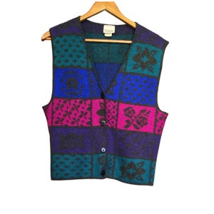 Può includere: Un gilet vintage senza maniche con un design patchwork. Il gilet presenta uno scollo a V e una chiusura con bottoni. La combinazione di colori include quadrati verde acqua, blu, viola e fucsia con motivi floreali e geometrici. Il gilet è esposto su una gruccia di legno.