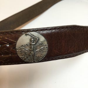 Może przedstawiać: Brązowy sk&oacute;rzany pasek ze srebrną klamrą z motywem golfa. Pasek posiada pleciony fragment i szczeg&oacute;łowy obraz golfisty wykonującego zamach kijem. Sk&oacute;ra wydaje się zużyta.
