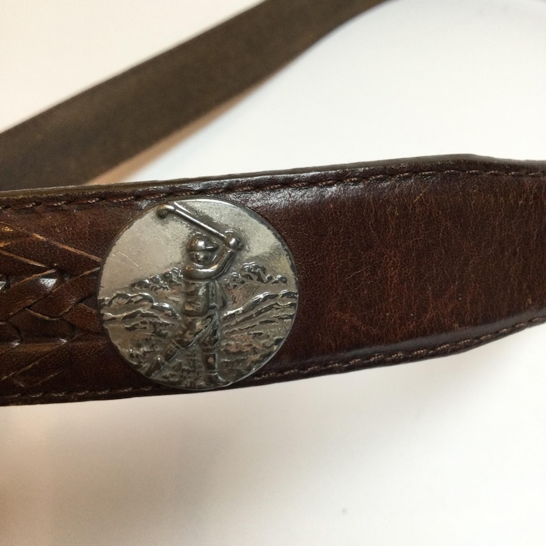 Może przedstawiać: Brązowy sk&oacute;rzany pasek ze srebrną klamrą z motywem golfa. Pasek posiada pleciony fragment i szczeg&oacute;łowy obraz golfisty wykonującego zamach kijem. Sk&oacute;ra wydaje się zużyta.