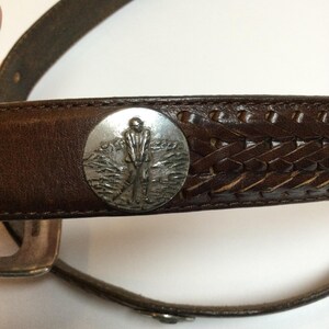 Może przedstawiać: Brązowy sk&oacute;rzany pasek ze srebrną okrągłą klamrą przedstawiającą golfistę. Pasek ma plecioną sk&oacute;rzaną sekcję i srebrną klamrę.