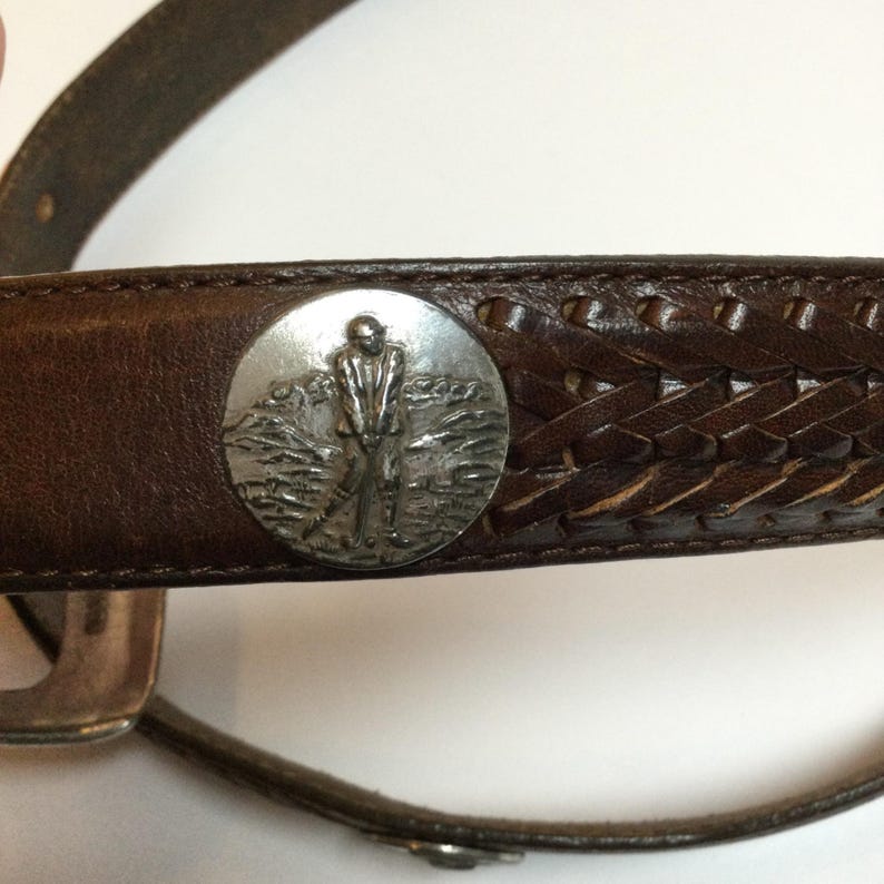 Może przedstawiać: Brązowy sk&oacute;rzany pasek ze srebrną okrągłą klamrą przedstawiającą golfistę. Pasek ma plecioną sk&oacute;rzaną sekcję i srebrną klamrę.