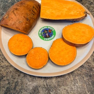 May include: An assortment of orange sweet potatoes, whole and sliced, arranged on a beige plate. The plate also features a green and white sticker with the text "Caribbean Spice Company". The sweet potatoes are a vibrant orange color.