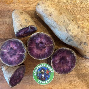 May include: An assortment of purple sweet potatoes, some whole and some cut to reveal the vibrant purple flesh. A Caribbean Spice Company sticker is also visible. The potatoes are on a wooden surface.
