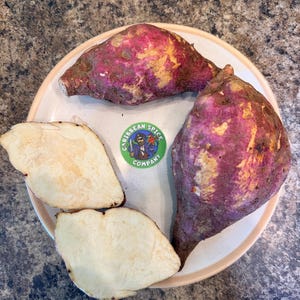 May include: A plate with two whole sweet potatoes and two sliced pieces. The sweet potatoes have a dark purple skin with yellow and brown patches. The sliced pieces are light beige. A green and white sticker with the text "Caribbean Spice Company" is also on the plate.