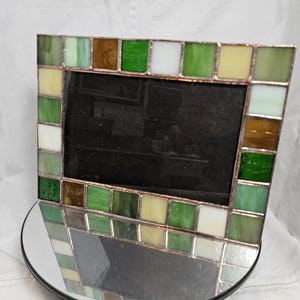 May include: A rectangular picture frame constructed from individual square pieces of stained glass in various shades of green, white, and brown. The glass tiles are joined together with thin copper-colored metal solder lines, creating a grid pattern.