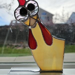 May include: A stained glass chicken figurine with large, black eyes and a red comb. The chicken is yellow with red accents and is standing on a silver base.