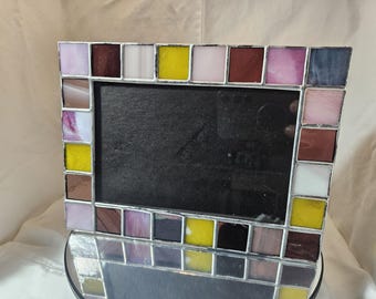 May include: A rectangular stained glass picture frame with a mosaic tile design. The frame features a variety of colors, including pink, yellow, brown, and white. The frame is designed to hold a photograph.