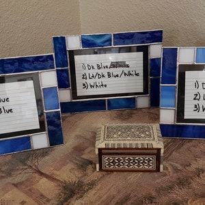 May include: Three stained glass picture frames with blue and white glass pieces. Each frame has a handwritten note listing the colors used in the frame. The note in the left frame reads: 1) Dark Blue 2) Cobalt Blue 5) White. The note in the center frame reads: 1) Dk Blue 2) Lt/Dk Blue/White 3) White. The note in the right frame reads: 1) Dark Blue/White 2) Light Blue/White 3) White.
