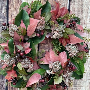 May include: A circular wreath featuring a mix of deep green magnolia leaves, dusty pink foliage, and clusters of small, light-colored dried flowers. Dark purple and burgundy berries are interspersed throughout the arrangement, creating a textured look.