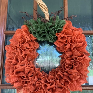 May include: A burlap wreath in the shape of a pumpkin with a brown stem and green burlap leaves. The wreath is decorated with brown curly ribbon.